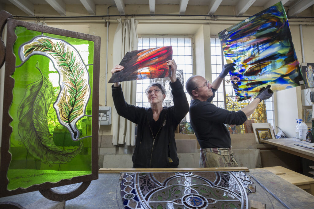Françoise Théallier et Philippe Riffaud levant les bras pour exposer à travers la lumière les échantillons de vitraux qu'ils tiennent dan leurs mains 